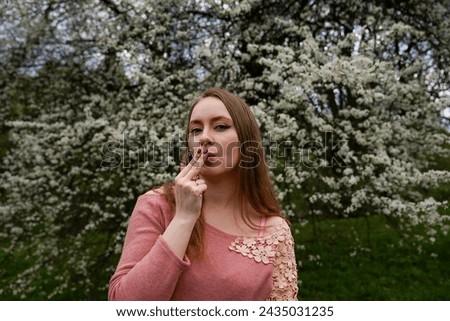 beautiful blond natural hair woman in pink outfit is posing in botanical garden park near blooming tree with flowers. Lady hold flower near mouth trying to taste and smell scent. Pretend to be smoking