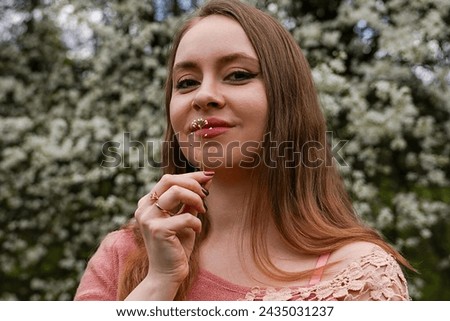 beautiful blond natural hair woman in pink outfit is posing in botanical garden park near blooming tree with flowers. Lady hold flower near mouth trying to taste and smell scent. Pretend to be smoking