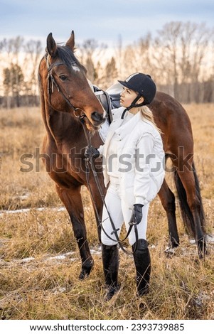 Beautiful blond professional female jockey standing near horse in field in winter. Friendship with horse. High quality photo