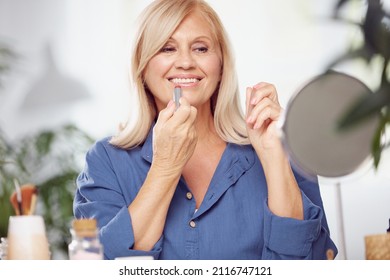 A beautiful blond senior woman sitting at home and putting lipstick on the lips. - Powered by Shutterstock