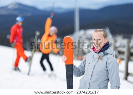Beautiful blond ski girl posing on the snow. Group of friends fooling in the background. Female skier smiling.
