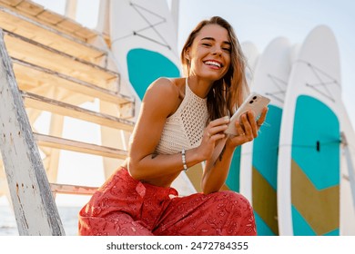 beautiful blond smiling woman posing on summer beach with surfboards on background in sunny weather dressed in stylish boho trend apparel knitted top and printed red trousers, using smart phone - Powered by Shutterstock