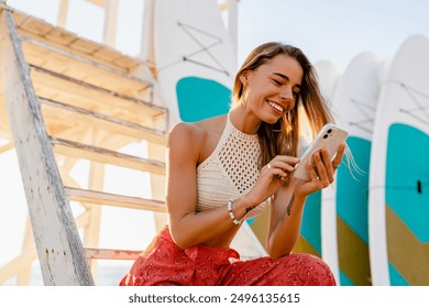 beautiful blond smiling woman posing on summer beach with surfboards on background in sunny weather dressed in stylish boho trend apparel knitted top and printed red trousers, using smart phone - Powered by Shutterstock