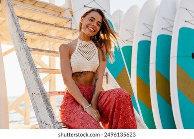 beautiful blond smiling woman posing on summer beach with surfboards on background in sunny weather dressed in stylish boho trend apparel knitted top and printed red trousers - Powered by Shutterstock