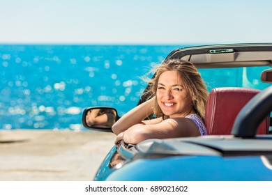 Beautiful blond smiling young woman in convertible top automobile looking sideways while parked near ocean waterfront - Powered by Shutterstock