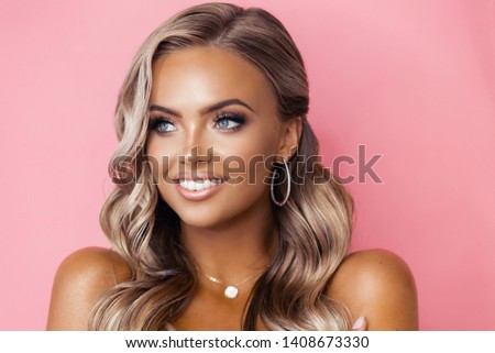 Beautiful blond tanned woman with wavy hair posing on pink backround wearing diamond accessories