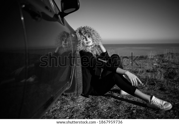 Beautiful blond\
woman with afro curly hair wearing black trouser suit and white\
sneakers sitting on the ground leaning on her car and enjoying\
sunshine. Black and white\
shot