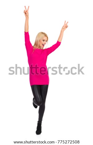 Beautiful blond woman in black leggings, high heels and pink sweater is standing on one leg, holding arms raised and shouting. Full length studio shot isolated on white.