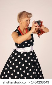 Beautiful blond woman in pinup style, dressed in a polka-dot dress, stands and holds a camera in front of her, looking in the wrong viewfinder, does not know how, is trained, white background.