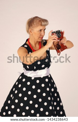 Beautiful blond woman in pinup style, dressed in a polka-dot dress, stands and holds a camera in front of her, looking in the wrong viewfinder, does not know how, is trained, white background.