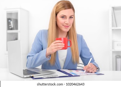 Beautiful blonde in a blue suit with a red cup in her hand at work. Young woman working on laptop.