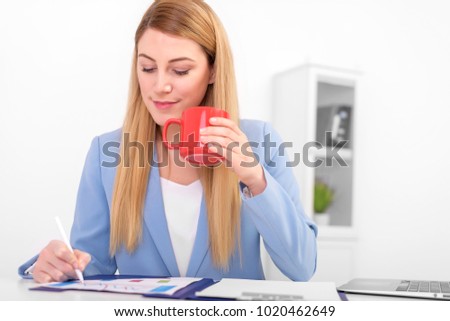 Beautiful blonde in a blue suit with a red cup in her hand at work. Young woman working on laptop.