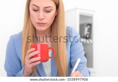 Beautiful blonde in a blue suit with a red cup in her hand at work. Young woman working on laptop.