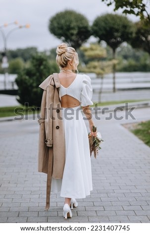 A beautiful blonde bride with a bouquet of wild flowers with reeds walks in autumn with a beige coat. Wedding photography, portrait.