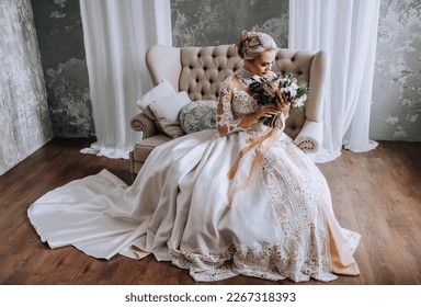 A beautiful blonde bride in a white lace dress sits on a beige sofa with a bouquet of flowers in a retro studio on a gray background. Wedding photography, portrait.