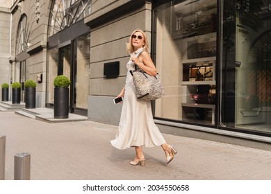 Beautiful Blonde Caucasian Mature Woman Walking While Shopping On The City Street, Carrying Handbag Outdoors. Middle-aged Businesswoman Buying Goods Clothes