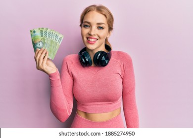 Beautiful blonde caucasian woman wearing gym clothes holding chilean pesos looking positive and happy standing and smiling with a confident smile showing teeth 