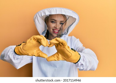 Beautiful blonde caucasian woman wearing protective beekeeper uniform smiling in love doing heart symbol shape with hands. romantic concept. 