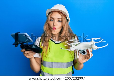 Beautiful blonde caucasian woman wearing safety hard using drone making fish face with mouth and squinting eyes, crazy and comical. 