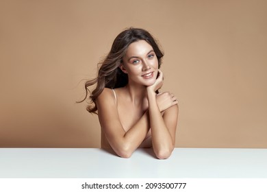 El hermoso pelo rubio y rizado en un traje de cuerpo beige está sentado en una mesa blanca  Perfecto maquillaje natural de cuerpo delgado  Cabello largo  cosméticos naturales
