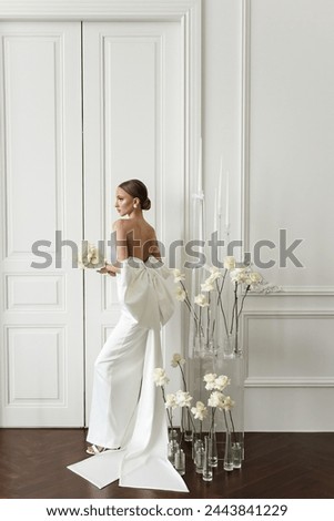 A beautiful blonde in an elegant white dress posing in a photo studio with a white classic interior and white roses.