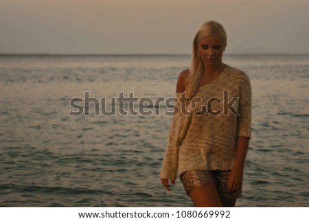 Beautiful blonde european model dressed beige romantic sweater looking down walking into the dark sea water on beach at sunset background. Summer time, vacation, travel and explore the world concept.
