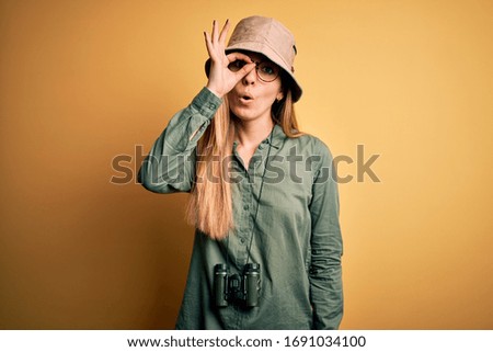 Beautiful blonde explorer woman with blue eyes wearing hat and glasses using binoculars doing ok gesture shocked with surprised face, eye looking through fingers. Unbelieving expression.