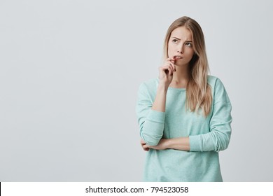 Beautiful blonde female with puzzled expression, keeps finger on lips, looks aside with bewilderment, poses against gray background. Body language and face expression