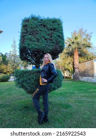 A Beautiful Blonde Girl In A Black Leather Jacket Poses At An Unusual Bush Against A Background Of Greenery