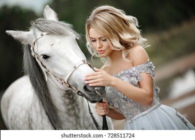 Beautiful blonde girl in dress strokes a gray horse on nature in summer