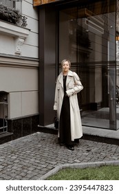 beautiful blonde girl dressed in beige oversized trench coat, black midi dress, loafers, bag, stylish fashion outfit, full length lifestyle model - Shutterstock ID 2395447823