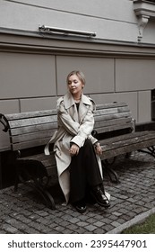 beautiful blonde girl dressed in beige oversized trench coat, black midi dress, loafers, bag, stylish fashion outfit, full length lifestyle model sitting - Shutterstock ID 2395447901