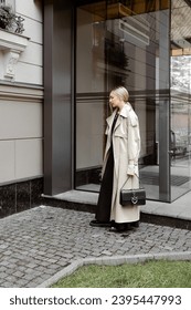 beautiful blonde girl dressed in beige oversized trench coat, black midi dress, loafers, bag, stylish fashion outfit, full length lifestyle model - Shutterstock ID 2395447993