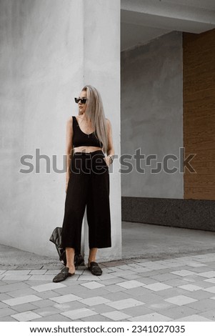 beautiful blonde girl dressed in black oversized culottes, top, mules, bag, sunglasses, accessories, stylish fashion outfit, full length lifestyle model