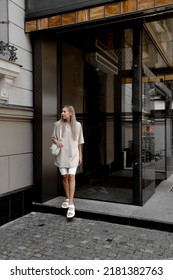 Beautiful Blonde Girl Dressed In White Bike Shorts, Beige Oversized T Shirt, White Bag And Sandals, Accessories, Stylish Trendy Summer Fashion Outlook, Full Height Lifestyle Model