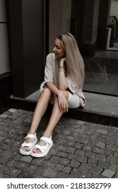 Beautiful Blonde Girl Dressed In White Bike Shorts, Beige Oversized T Shirt, White Bag And Sandals, Accessories, Stylish Trendy Summer Fashion Outlook, Lifestyle Model Sitting