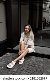 Beautiful Blonde Girl Dressed In White Bike Shorts, Beige Oversized T Shirt, White Bag And Sandals, Accessories, Stylish Trendy Summer Fashion Outlook, Lifestyle Model Sitting