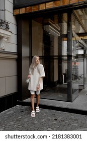 Beautiful Blonde Girl Dressed In White Bike Shorts, Beige Oversized T Shirt, White Bag And Sandals, Accessories, Stylish Trendy Summer Fashion Outlook, Full Height Lifestyle Model