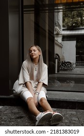 Beautiful Blonde Girl Dressed In White Bike Shorts, Beige Oversized T Shirt, White Bag And Sandals, Accessories, Stylish Trendy Summer Fashion Outlook, Lifestyle Model Sitting