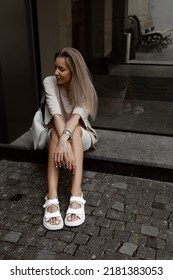 Beautiful Blonde Girl Dressed In White Bike Shorts, Beige Oversized T Shirt, White Bag And Sandals, Accessories, Stylish Trendy Summer Fashion Outlook, Lifestyle Model Sitting