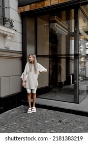 Beautiful Blonde Girl Dressed In White Bike Shorts, Beige Oversized T Shirt, White Bag And Sandals, Accessories, Stylish Trendy Summer Fashion Outlook, Full Height Lifestyle Model