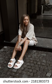 Beautiful Blonde Girl Dressed In White Bike Shorts, Beige Oversized T Shirt, White Bag And Sandals, Accessories, Stylish Trendy Summer Fashion Outlook, Lifestyle Model Sitting