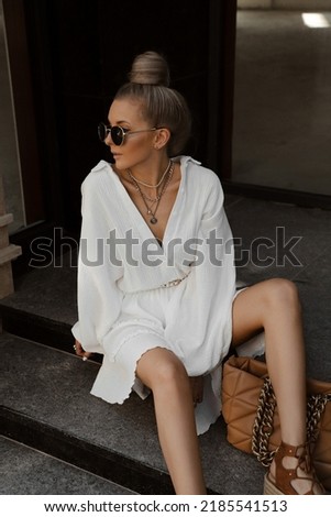 beautiful blonde girl dressed in white oversized magnificent dress, brown lace-up sandals and bag, accessories, sunglasses, bundle hairstyle, stylish summer fashion outlook, lifestyle model sitting
