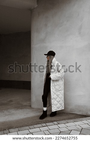 beautiful blonde girl dressed in white oversized long jacket, black leggings and sweater, boots, cap, bag in hands, stylish fashion winter outfit, full length lifestyle model