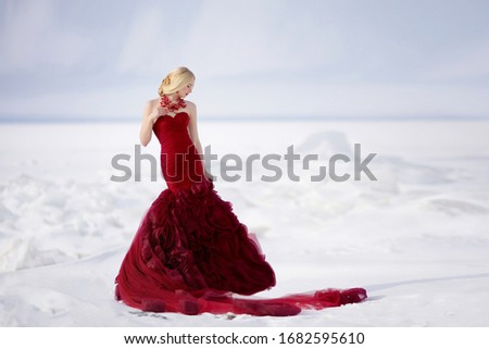a beautiful blonde girl with a beautiful hairstyle in a long red dress stands on the ice of the frozen sea