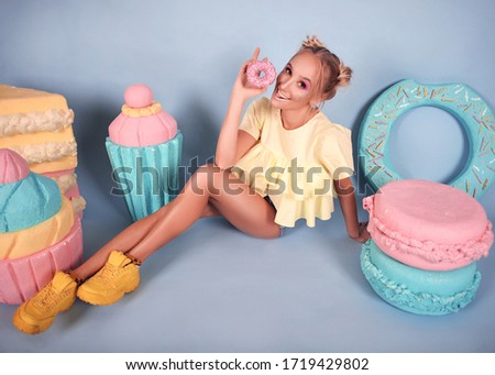 beautiful blonde girl have fun and fool around with big cakes and macarons and donuts on a blue background with bright makeup