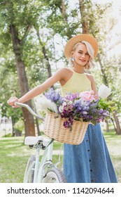beautiful blonde girl holding bike with basket of flowers in park