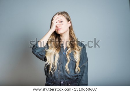 Beautiful blonde girl peeps through hands, isolated on gray background