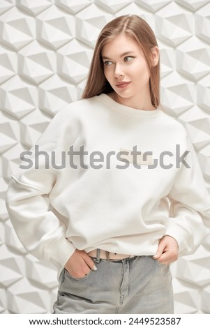 Beautiful blonde girl posing in a loose white jumper and wide jeans on a white geometric studio background. Women's fashion. Oversized style.