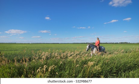 Beautiful blonde girl riding a horse at a countryside. Charming, sexy woman wearing a red dress riding a horse at a green field. Freedom, happiness, summer